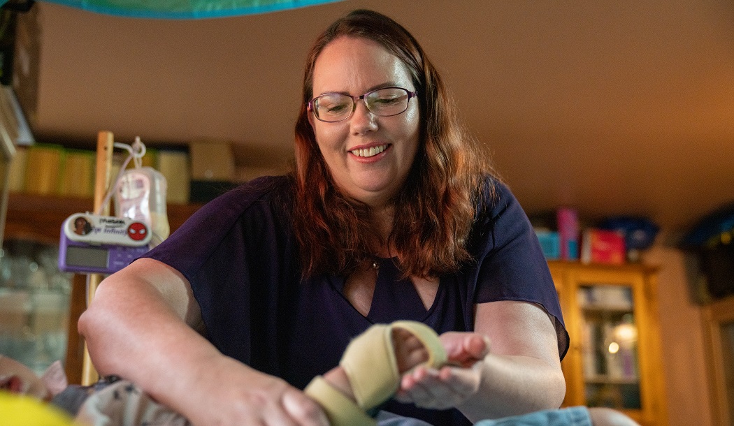 Debbie wears a purple shirt, reading glasses and has long reddish-brown hair below her shoulders. She is smiling while caring for a child with high needs. She is holding the child's hand and the child cannot be identified.
